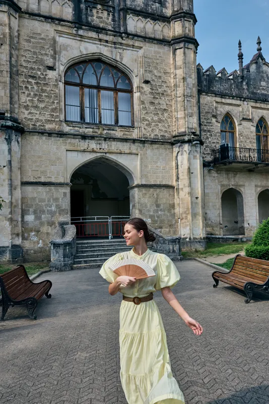 A woman in a pale yellow dress walks in a historic castle courtyard, holding a decorative fan as sunlight highlights the ancient stone architecture and tranquil surroundings.