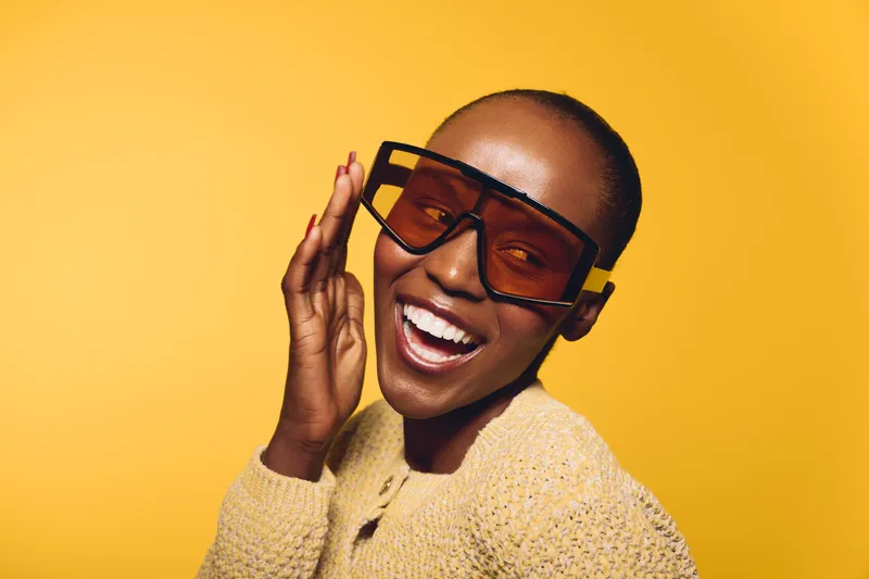 Happy black woman with dark skin wearing large stylish sunglasses and knitted sweater smiles joyfully on yellow background. Concept of fashion, beauty, and positive mood.