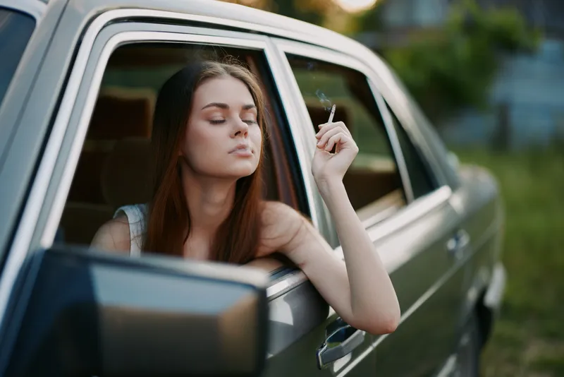 Young woman relaxing in car, smoking a cigarette, showcasing a carefree attitude in a natural green setting with warm evening light.