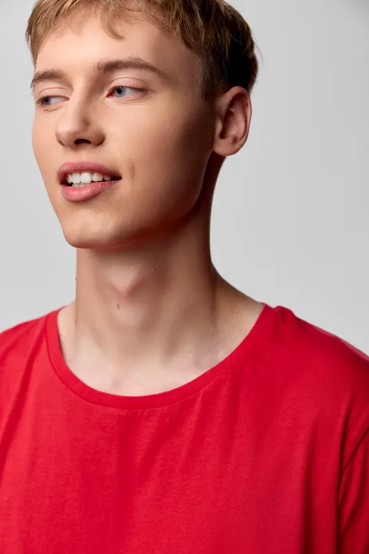 Young man in a casual red shirt poses in studio lighting with a soft smile, showcasing relaxed fashion and contemporary style against a clean background and natural expression.