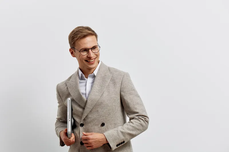 Modern professional young man with glasses in business casual attire holding a laptop and documents look away smiling confident pose on plain white background for corporate and workplace concept