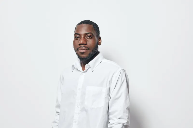 African American man with dark skin wearing white shirt standing against white background looking at camera with confident expression and slight smile in studio portrait.