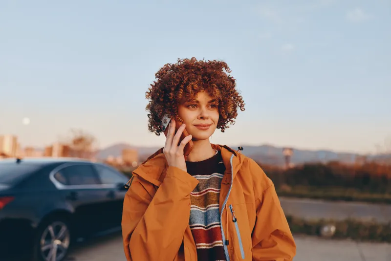 Woman lifestyle portrait outdoors at sunset, wearing orange jacket and striped sweater, curly hair framing her face, hand near cheek thoughtful gesture, road with cars in soft blur, warm calm