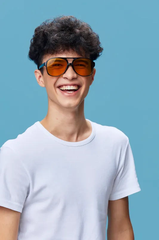 smiling man wearing orange sunglasses and white t-shirt with curly hair on blue background expressing happiness and youthfulness in casual style