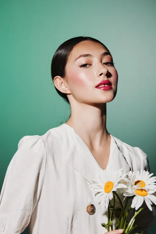 Elegant woman with shiny skin and bright lipstick poses holding white daisies, wearing a white outfit. Studio shot with soft light on a pastel green backdrop. Fashion and beauty concept.