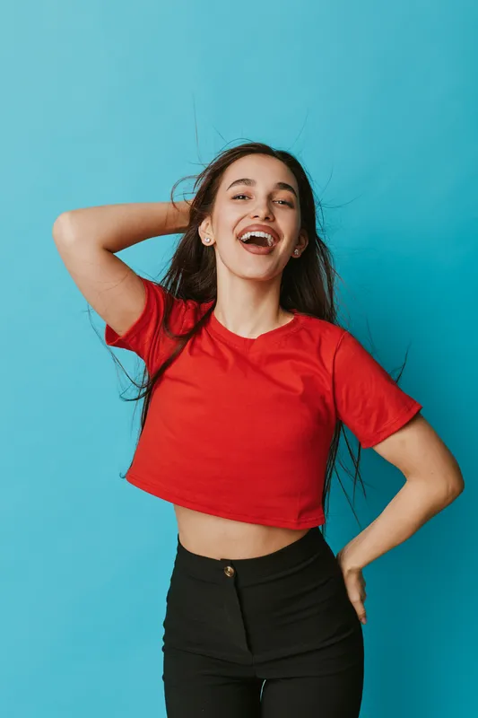 Young woman in red crop top and black pants posing with hand behind head and smiling against blue background. Casual style, happy expression and confident pose in studio.