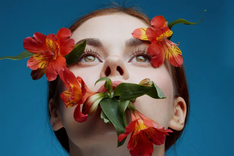 A young woman with red flowers around her face, expressing serenity against a blue background, highlighting beauty and nature