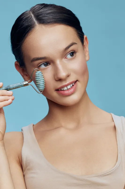 Young woman using a jade roller for skin care, smiling happily against a light blue background, promoting a healthy beauty routine or relaxation