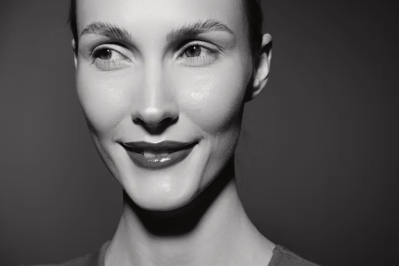 Close-up of a young white woman with fair skin and short hair smiling subtly against a dark background. Concept of natural beauty, facial expression, and monochrome portrait.