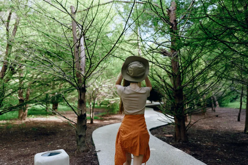 Woman hat walking path park trees nature back in summer scene, casual dress and wide brim hat lifted while strolling along winding paved trail surrounded by green woodland calm