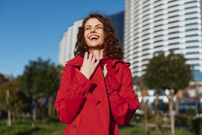 Young woman with curly hair laughs joyfully outdoors wearing a bright red coat against a cityscape background on a sunny day.
