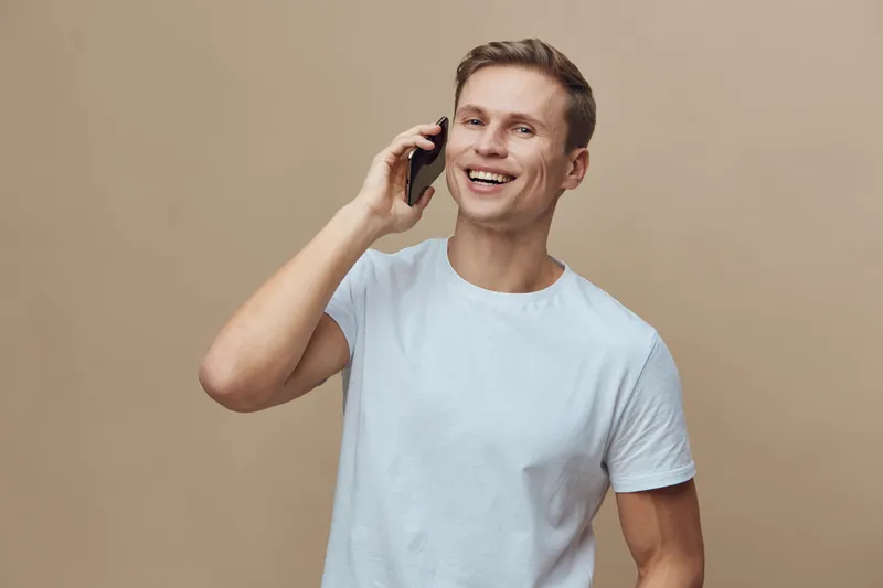 Smiling man talking on mobile phone, casual white t-shirt, cheerful expression, indoor studio shot, beige background, lifestyle concept, modern communication, happiness, leisure.