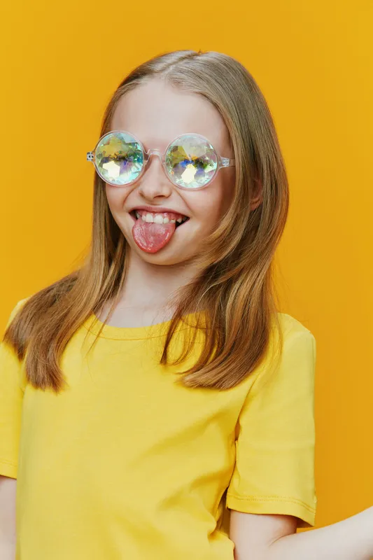 Young girl with shiny oversized glasses making a playful expression against a bright yellow background, wearing a vibrant yellow t shirt, conveying joy and summer vibes
