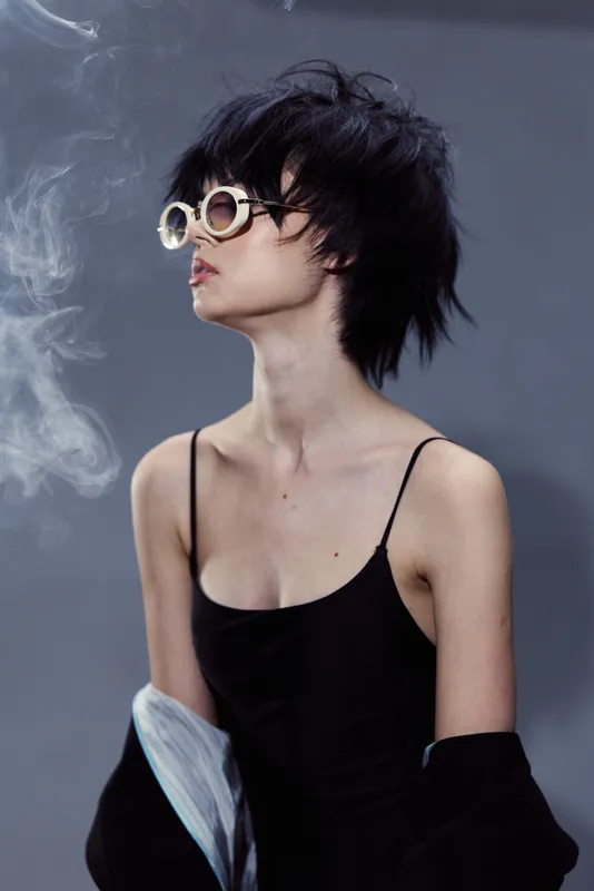 Stylish woman with short hair wearing sunglasses and black dress with smoke in background