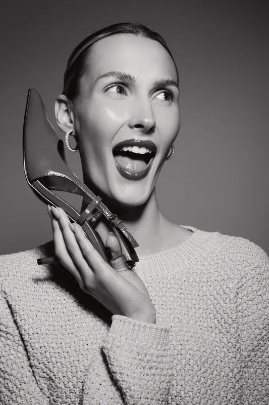 Young white woman with fair skin holds a high heel shoe near her face, smiling widely in black and white studio portrait. Concept for fashion and joy.