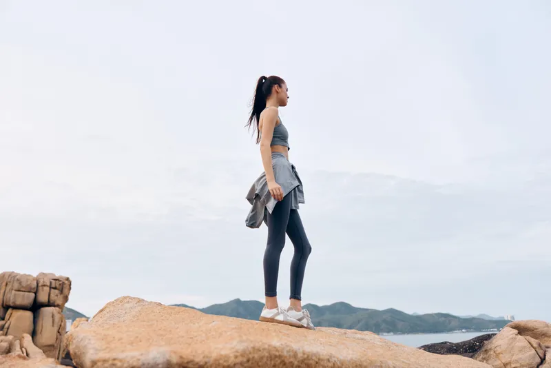 Young Asian woman in sportswear standing on a rock, showcasing fitness and determination against a coastal background with a serene atmosphere