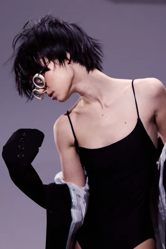 Stylish woman in black bodysuit and glasses posing with dynamic short hair and modern attitude indoors.