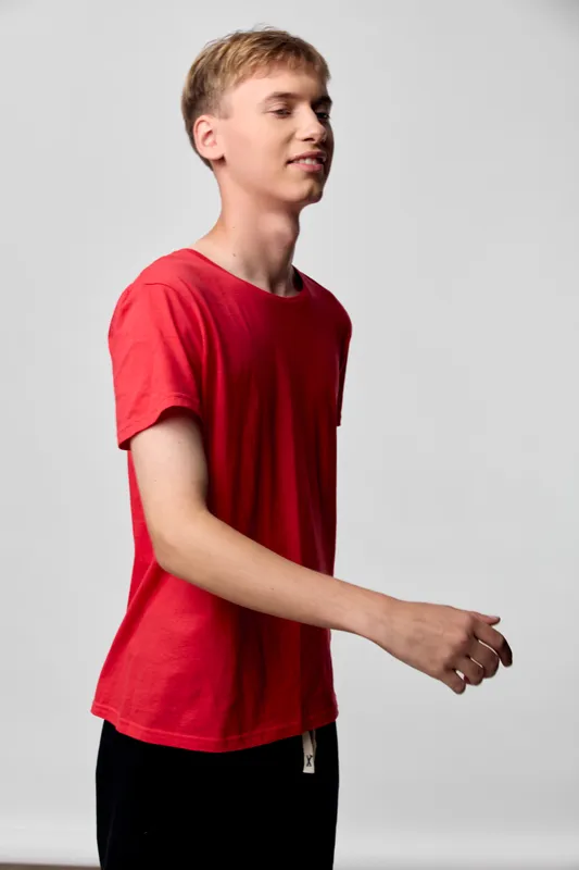 Young man in a red t shirt stands in a studio, captured in profile with relaxed posture and subtle movement, minimal background and clean lighting.