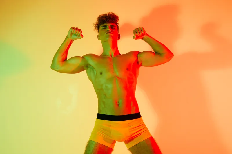 Muscular young man posing shirtless with flexed arms and defined abs in colorful lighting for fitness, strength, health and bodybuilder concept in studio background.