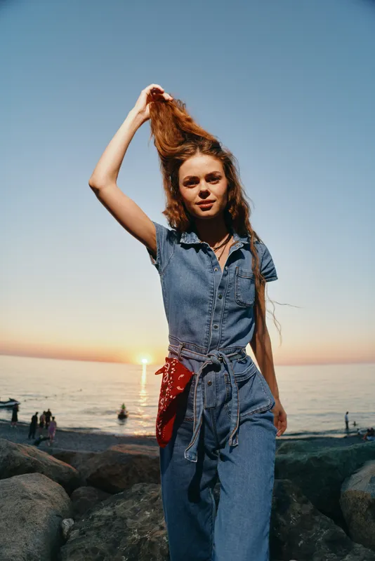 young woman in denim jumpsuit standing on rocky beach at sunset with calm sea and warm sunlight in background vibrant casual style outdoors
