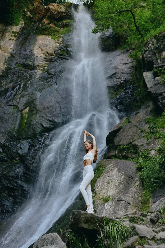 woman practicing yoga next to a tall waterfall, wearing white sportswear, surrounded by rocky cliffs and green forest, peaceful outdoor environment