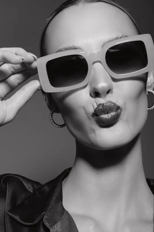 Young white woman with pale skin and slicked back hair wears large sunglasses and hoop earrings, making a playful kiss face in black and white portrait.