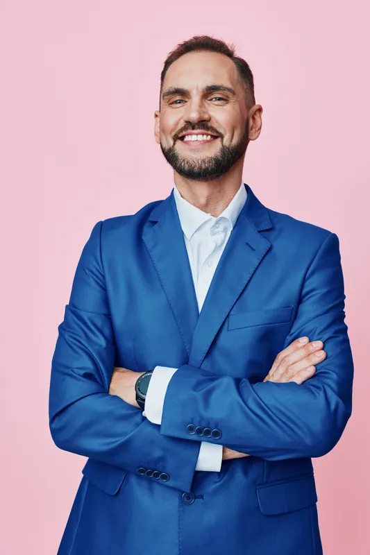 A confident male business professional stands with folded arms against a solid pink background, exuding positive energy, calm focus, and approachable determination.