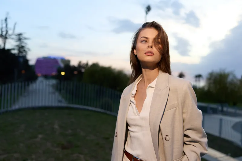 Confident business woman outdoors, dressed in a light blazer and blouse, showcasing elegance and professionalism against a serene sunset background.