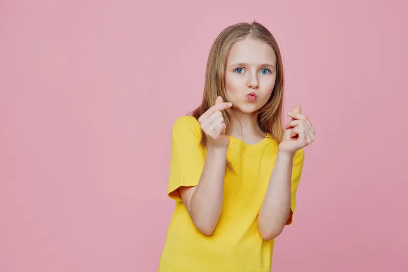 Young girl in a bright yellow shirt making a heart gesture against a pink background, showcasing joy and playfulness, ideal for cheerful themes