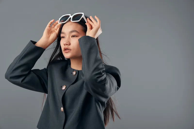 Young woman with long black hair wearing black coat and white sunglasses adjusting eyewear on head against gray studio background for fashion portrait and style concept.