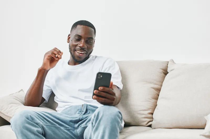 Happy young Black man enjoying conversation on smartphone at home, wearing casual white t shirt and jeans, relaxed atmosphere