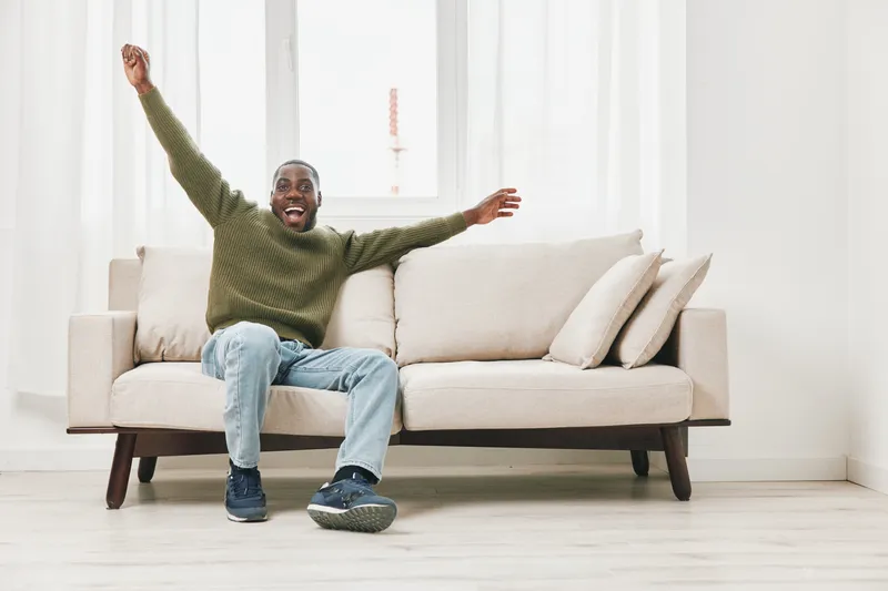 Excited young man celebrating joyfully on a comfortable couch in a bright, modern living room, conveying happiness and relaxation in a cozy atmosphere