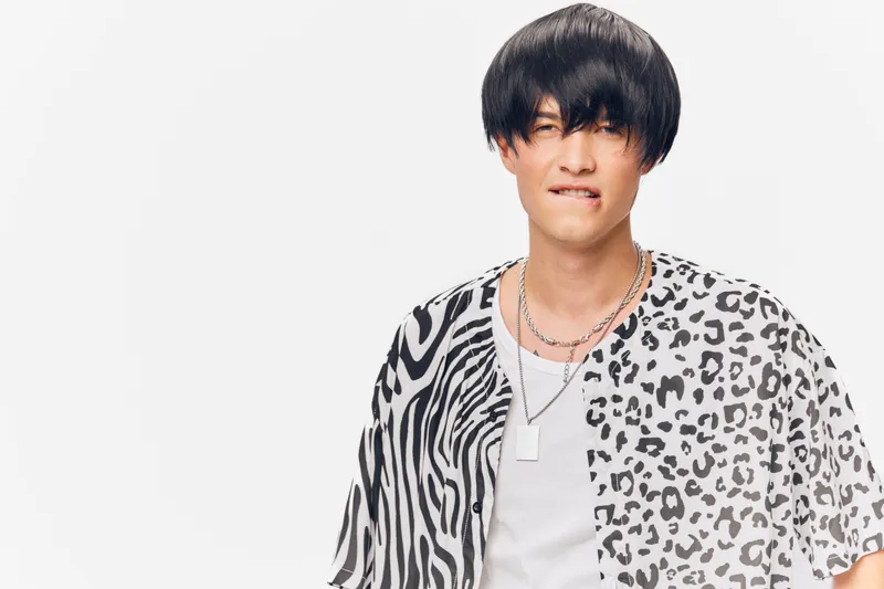 young man wearing a patterned shirt biting his lip with a playful expression, short black hair, silver necklaces, isolated on white background