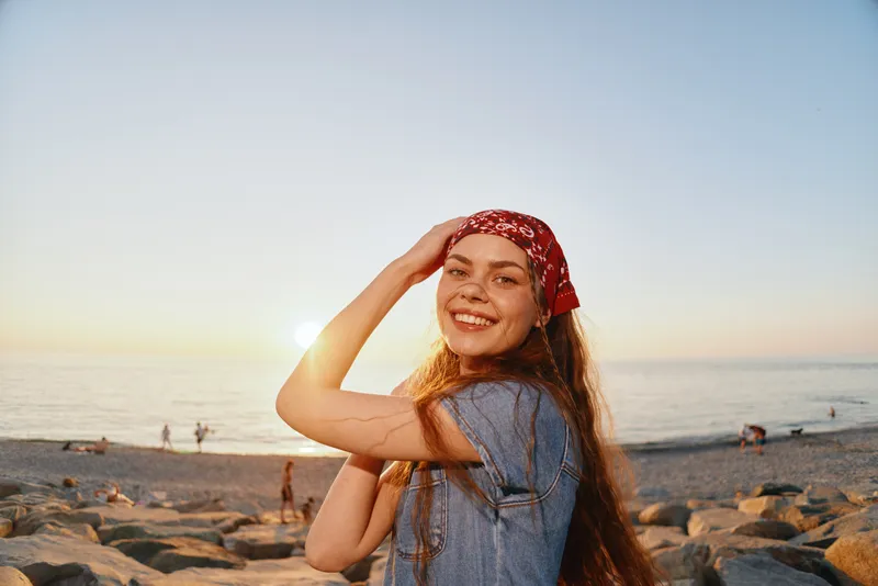 smiling woman with red headscarf enjoys sunset on rocky beach with calm sea and warm sunlight, feeling relaxed and happy outdoors