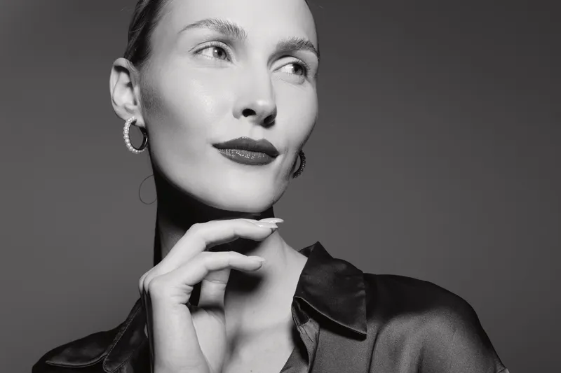A young Caucasian woman with fair skin poses thoughtfully, wearing hoop earrings and a dark satin blouse. The black and white portrait highlights elegance and classic beauty.