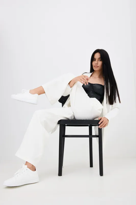 stylish woman sitting on black chair wearing white pants, black top, and white sneakers in minimalist white studio with long straight hair