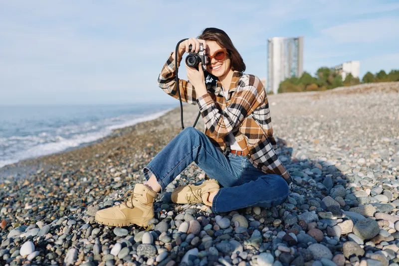 Photographer sitting on a pebble beach with camera, wearing plaid shirt, jeans and boots, capturing the sea and distant buildings in the background.