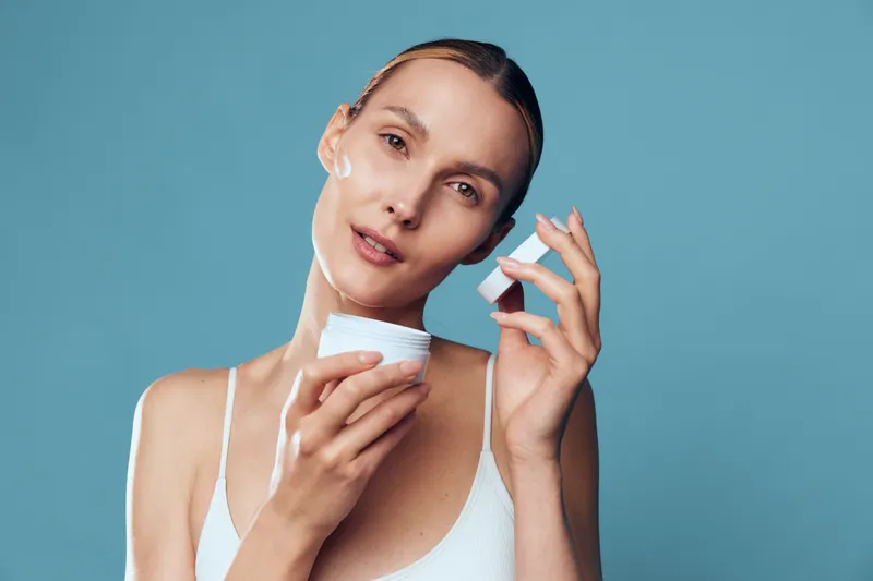 Young woman applies moisturizing cream on face holds white jar with skincare product has natural makeup and wet hair isolated on blue background in beauty and wellness concept.