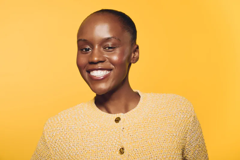 Smiling African woman with dark skin wearing a yellow knitted sweater stands against a yellow background. The image represents happiness, confidence, and natural beauty in portrait photography.