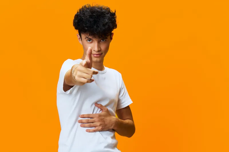 Young man with curly hair pointing forward confidently, wearing a white t-shirt against bright orange background, showcasing playful and energetic expression with casual style.