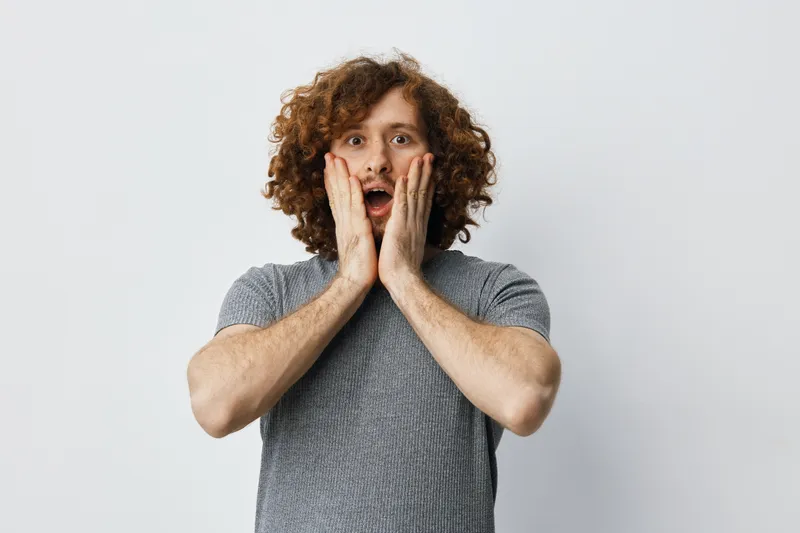 Surprised young man with curly hair expressing shock while touching his face against a neutral background, showcasing emotions and natural beauty