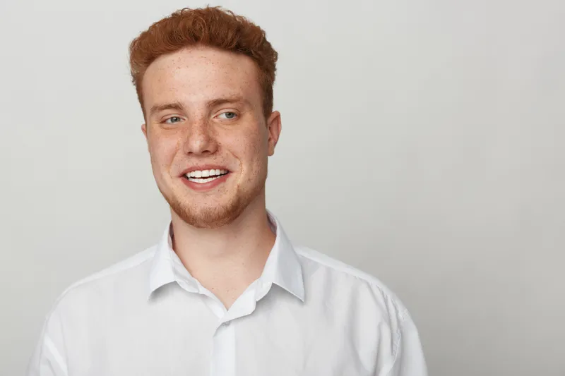 Smiling young man with red hair and beard wearing white shirt looks away on light background. Casual portrait of happy male model with natural expression and clean studio lighting.