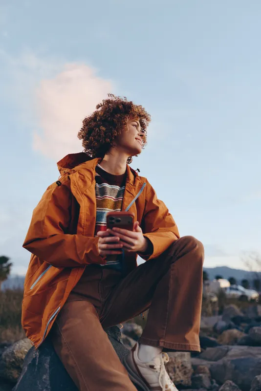 Woman lifestyle outdoor portrait, curly hair girl in orange jacket, sitting on rocks in nature landscape, holding smartphone and looking at screen, calm relaxed travel mood, blue sky