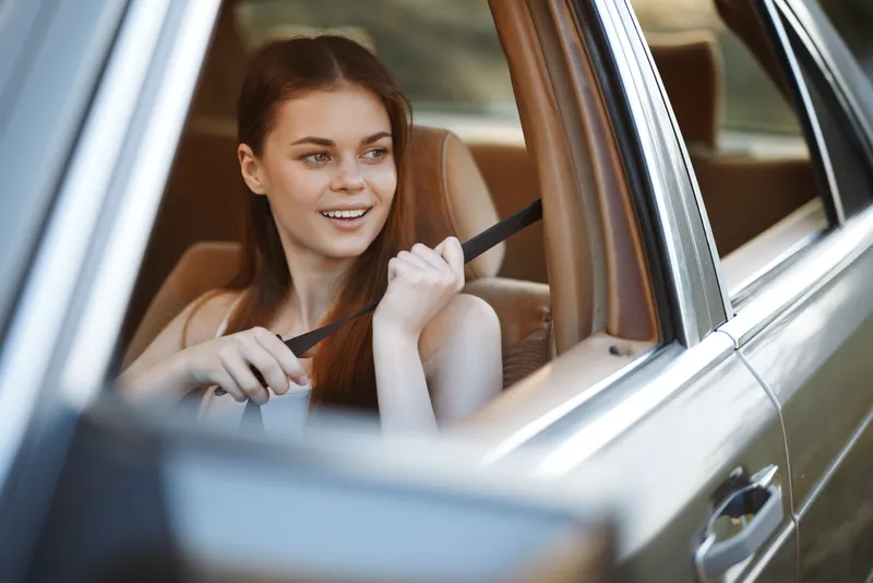 Young woman smiling while fastening seatbelt in a car, enjoying a safe road trip in a sunny outdoor setting with natural light.