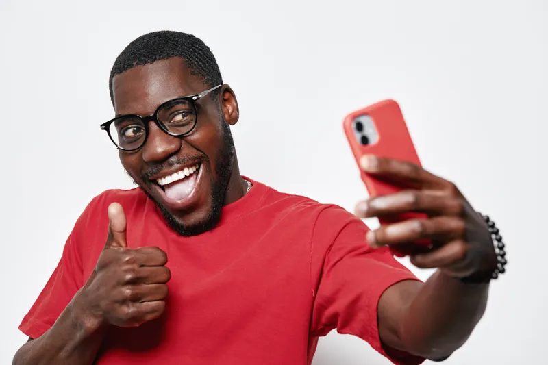 African American man with dark skin wearing glasses and red shirt taking selfie with smartphone smiling and showing thumbs up gesture on white background.