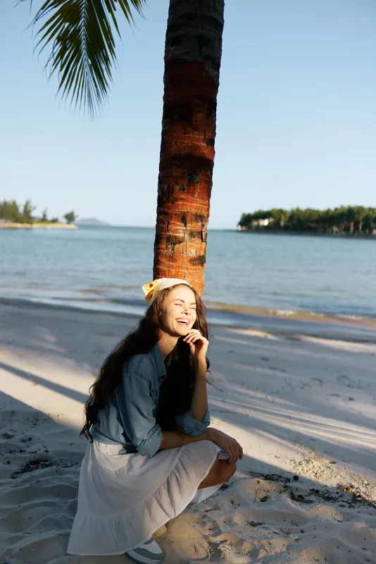 Woman sits under a palm tree on a sunny beach, wearing a white dress and denim shirt, calm expression, warm light, tranquil shoreline, tropical vibe.
