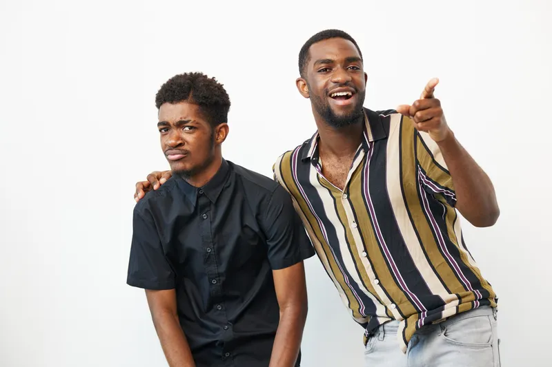 Two African American men enjoying a fun moment together, one smiling broadly while the other feigns annoyance. Expressive and lighthearted atmosphere in a neutral background.