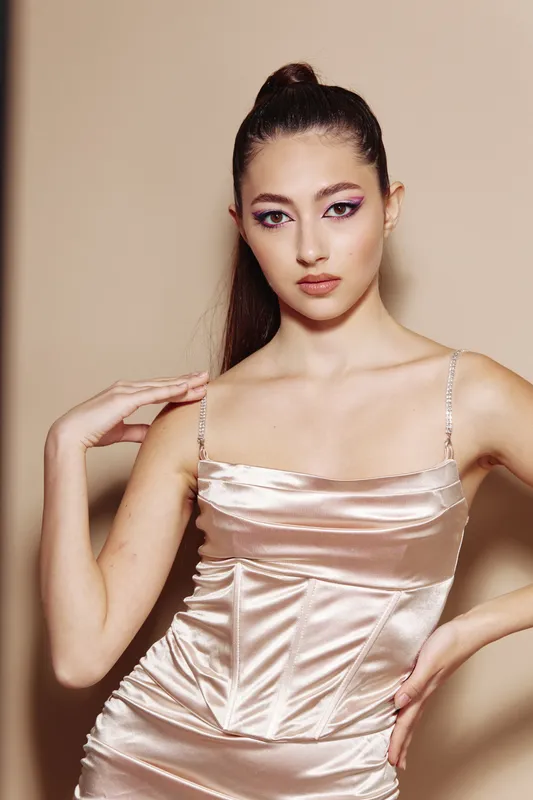 Young woman in a satin dress poses confidently against beige background with sharp makeup and sleek hairstyle. The image conveys beauty, fashion, and creative elegance.