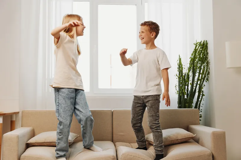 Two happy children jump on a beige sofa in a bright living room with natural light from the window and a green plant on the side. Concept of childhood fun and home activity.