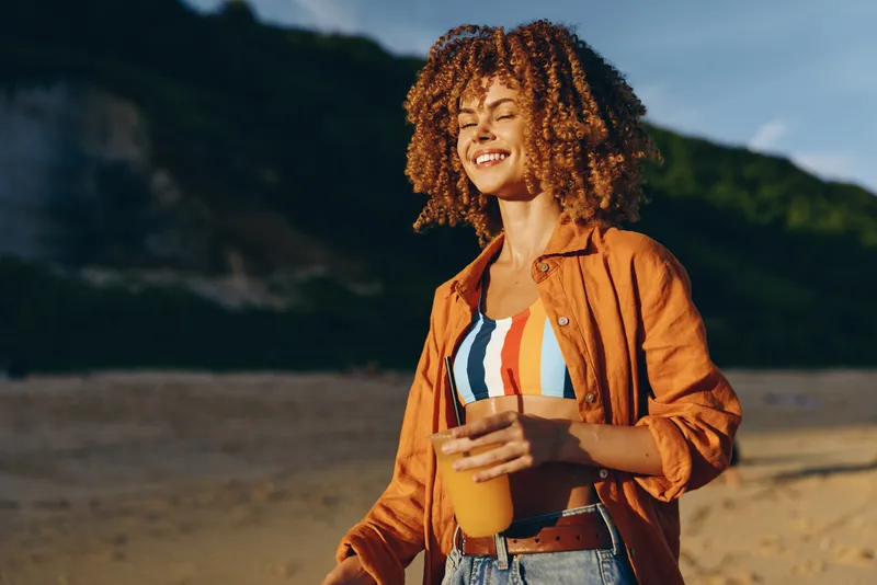 Young African American woman with curly hair enjoys fresh juice outdoors on a sunny day wearing a colorful striped top and orange shirt with a joyful smile.